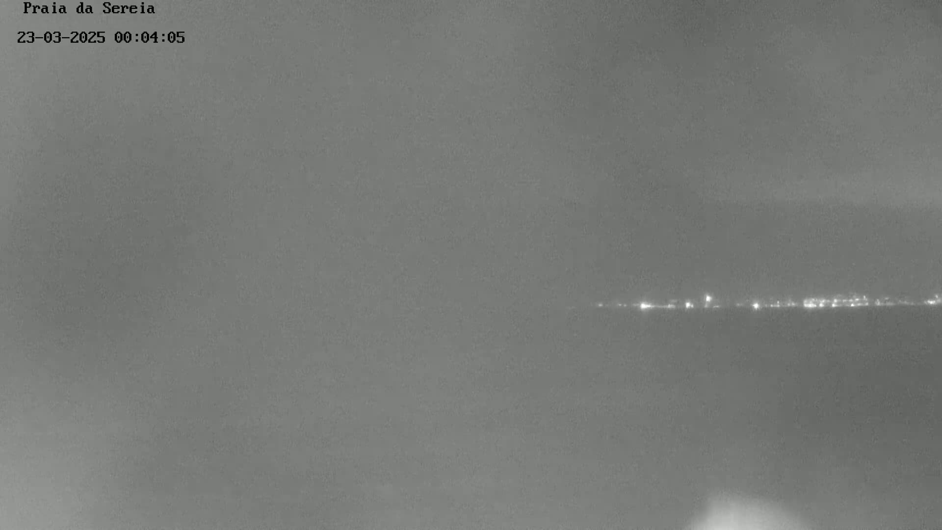 A long-distance nighttime shot shows a faint line of lights on the horizon over a dark body of water under a cloudy sky.