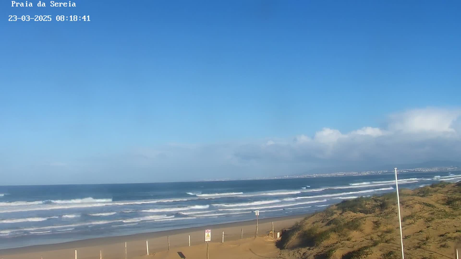 A sandy beach with small waves under a mostly clear blue sky with some scattered clouds is shown.