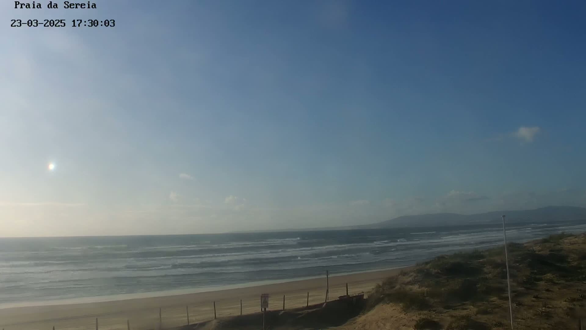 A mostly sunny day at a beach with a sandy shore, gentle waves, and distant mountains.