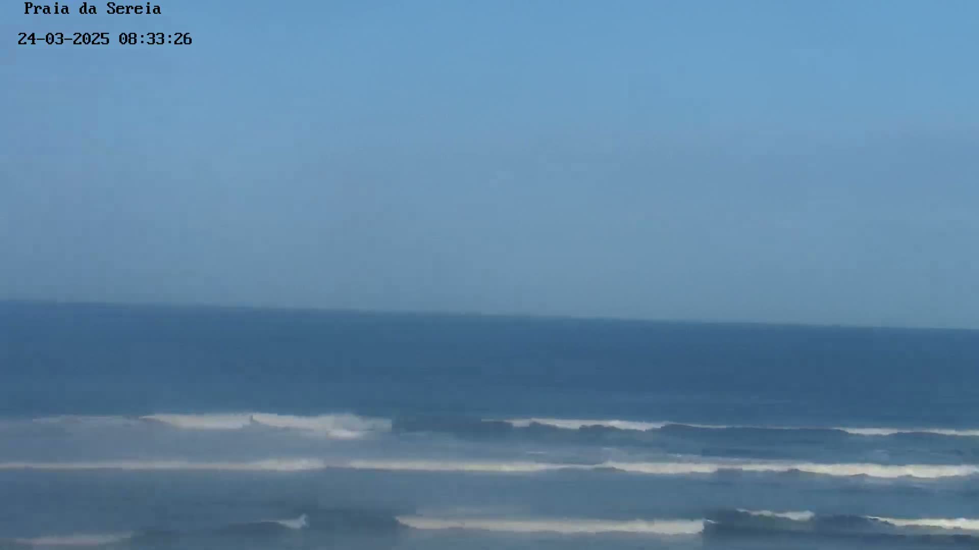A mostly clear blue sky overlooks a calm ocean with small waves breaking near the shore.