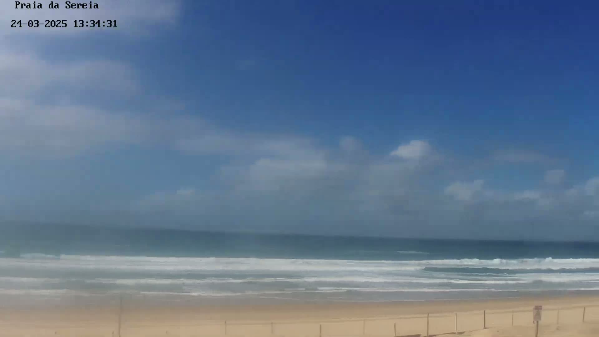 A mostly sunny day shows a sandy beach with gentle waves rolling into shore under a partly cloudy sky.