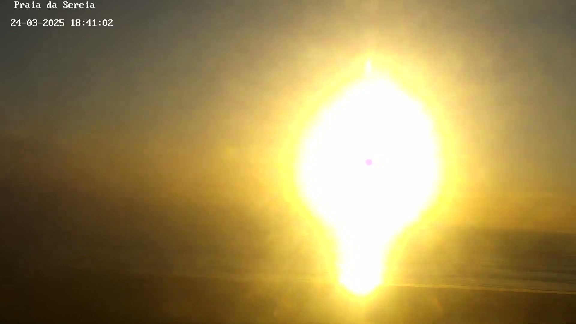 A hazy, bright yellow sun, appearing distorted, sets over a dark beach.