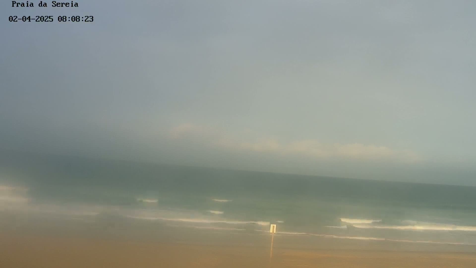 A hazy, overcast day shows a sandy beach and a calm ocean with small waves.