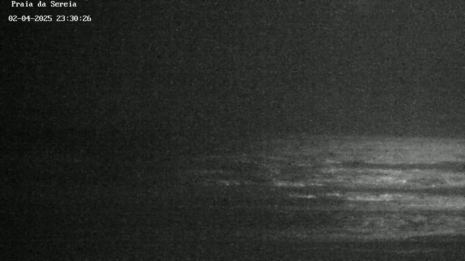A low-light nighttime image shows a dark, possibly sandy, beach or shoreline with subtle wave patterns visible in the low-light conditions.