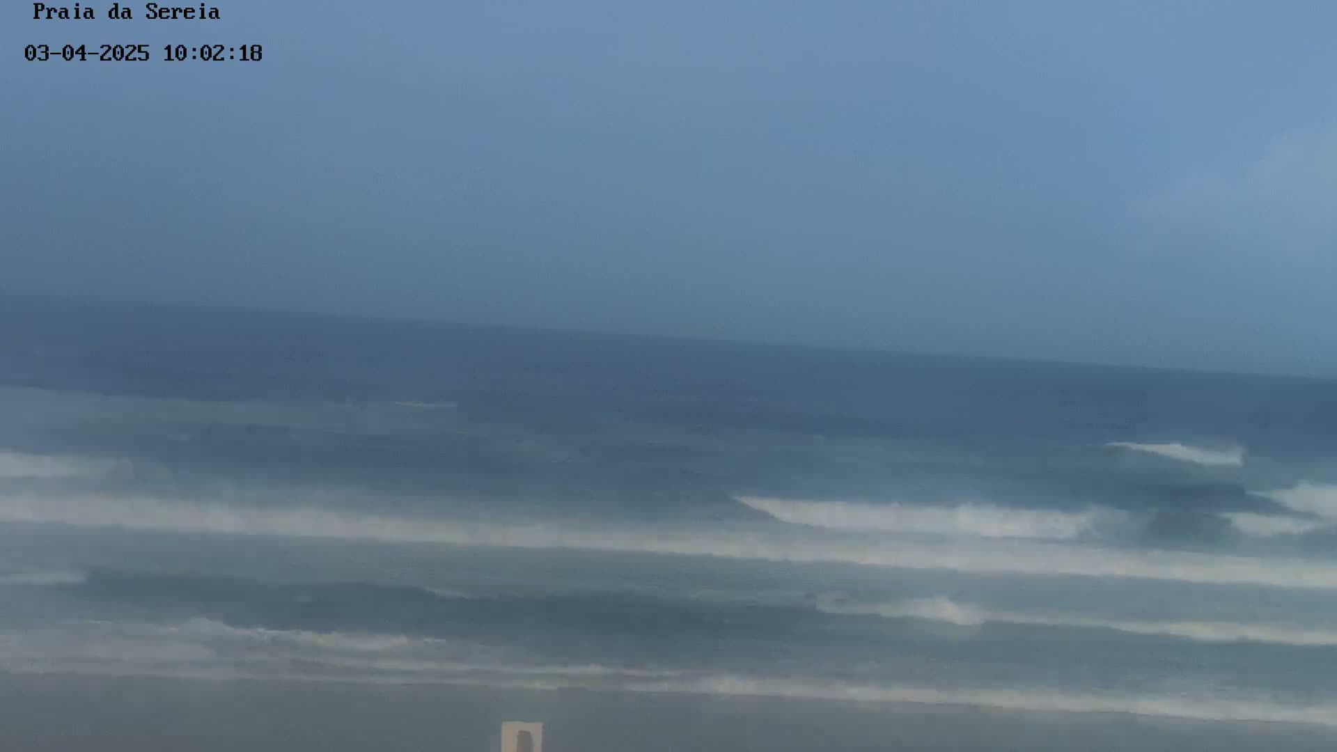 A hazy, overcast sky overlooks a calm ocean with small waves breaking on a beach.