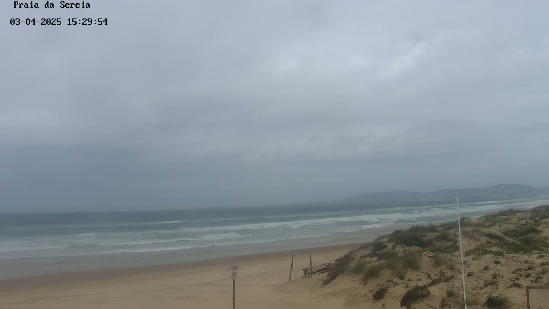 A sandy beach with rolling waves under an overcast sky stretches to a distant coastline.