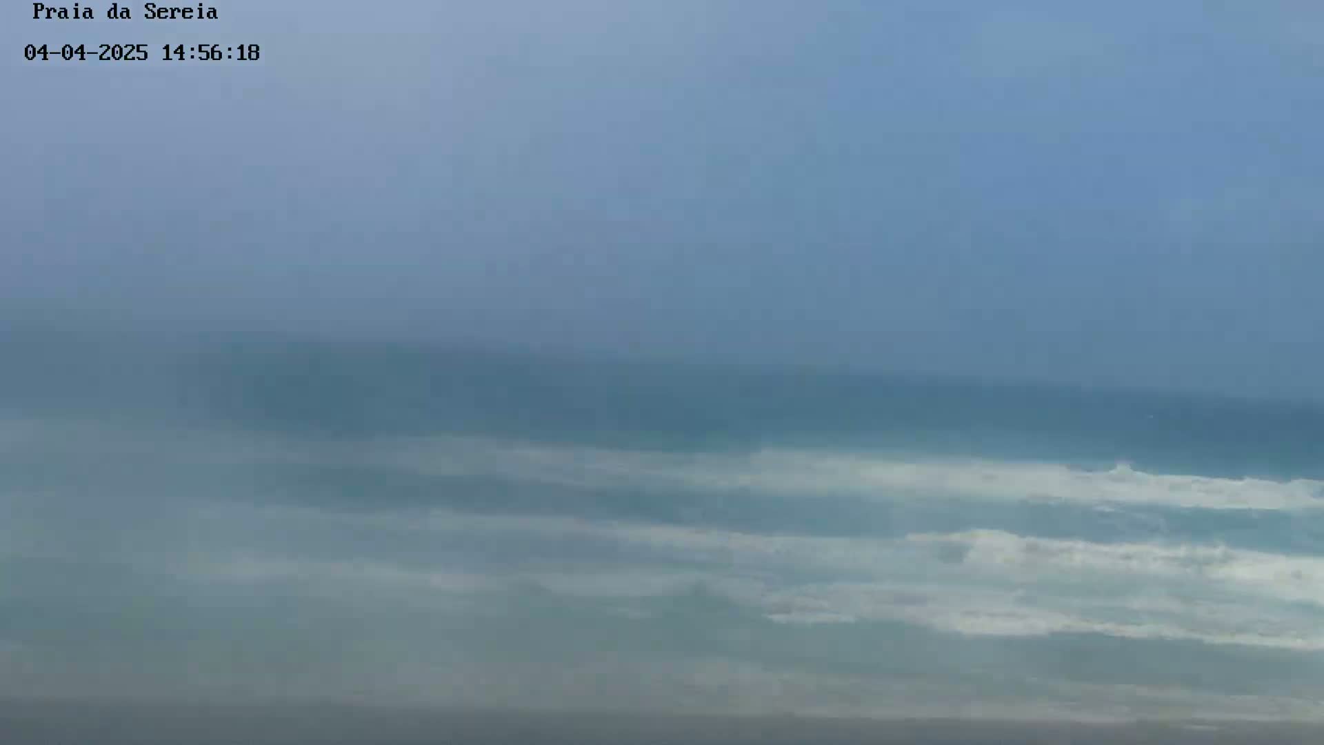 A hazy, overcast sky overlooks a calm ocean with small waves breaking gently near the shore.