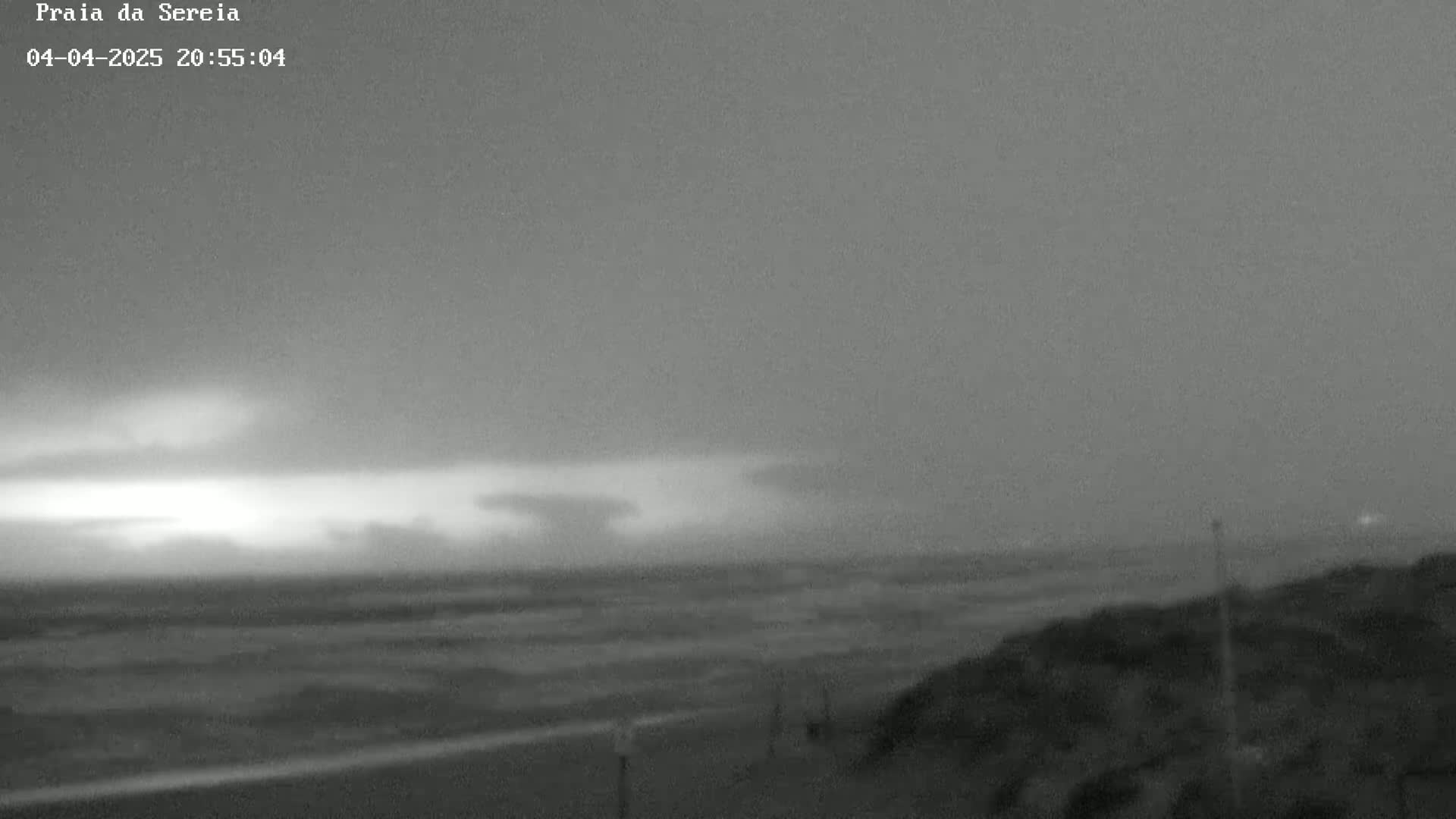 A nighttime, overcast beach scene shows a dark, wavy ocean under a stormy sky with faint lightning in the distance.