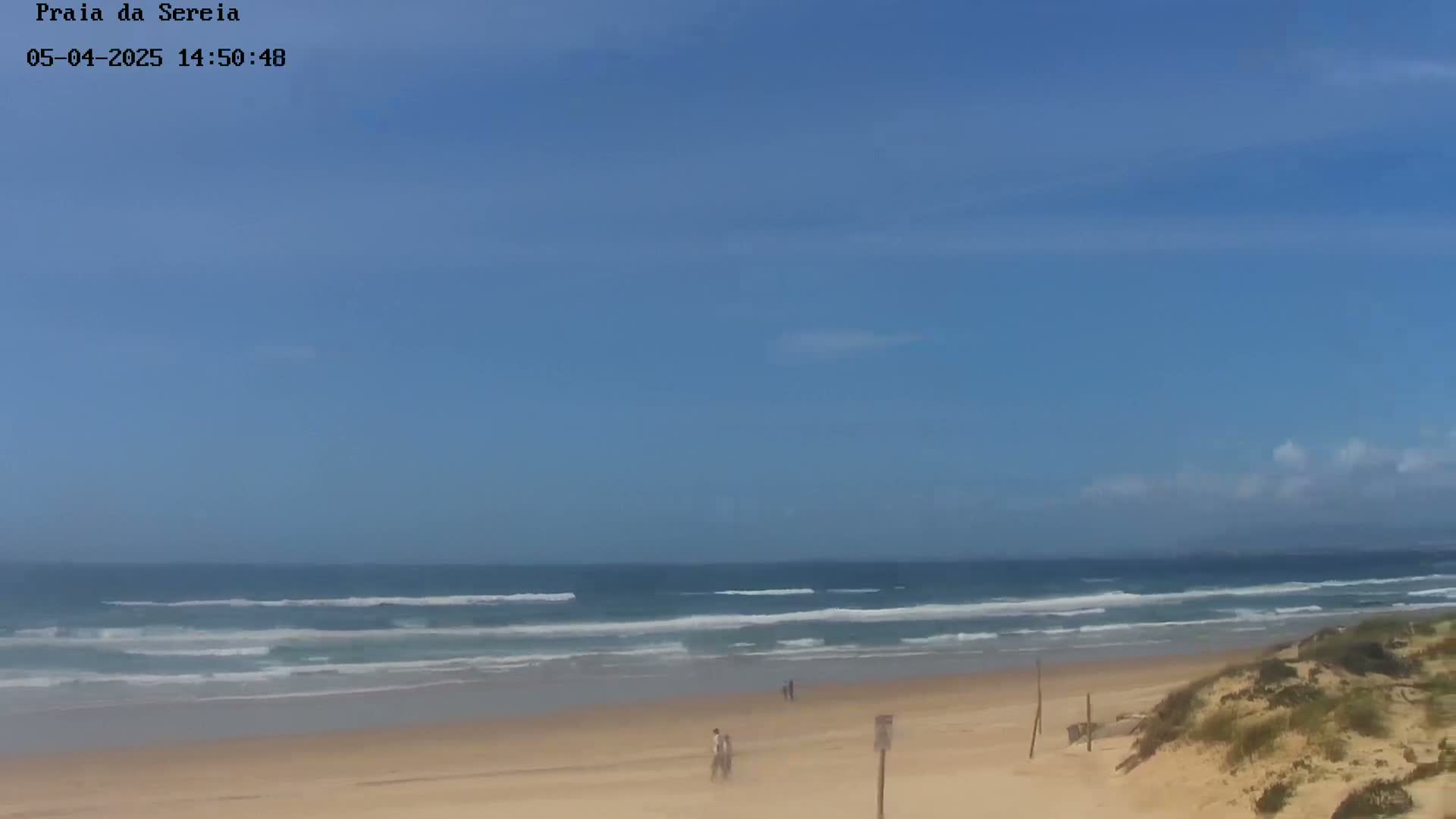 A sandy beach with gentle waves under a mostly clear, light blue sky.