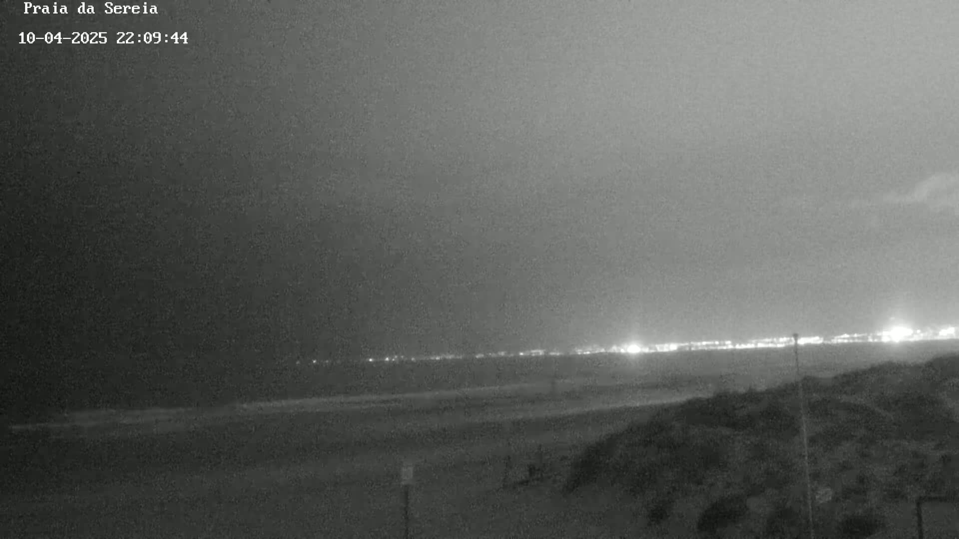 A dark, nighttime beach scene shows a distant line of lights across the water under an overcast sky.