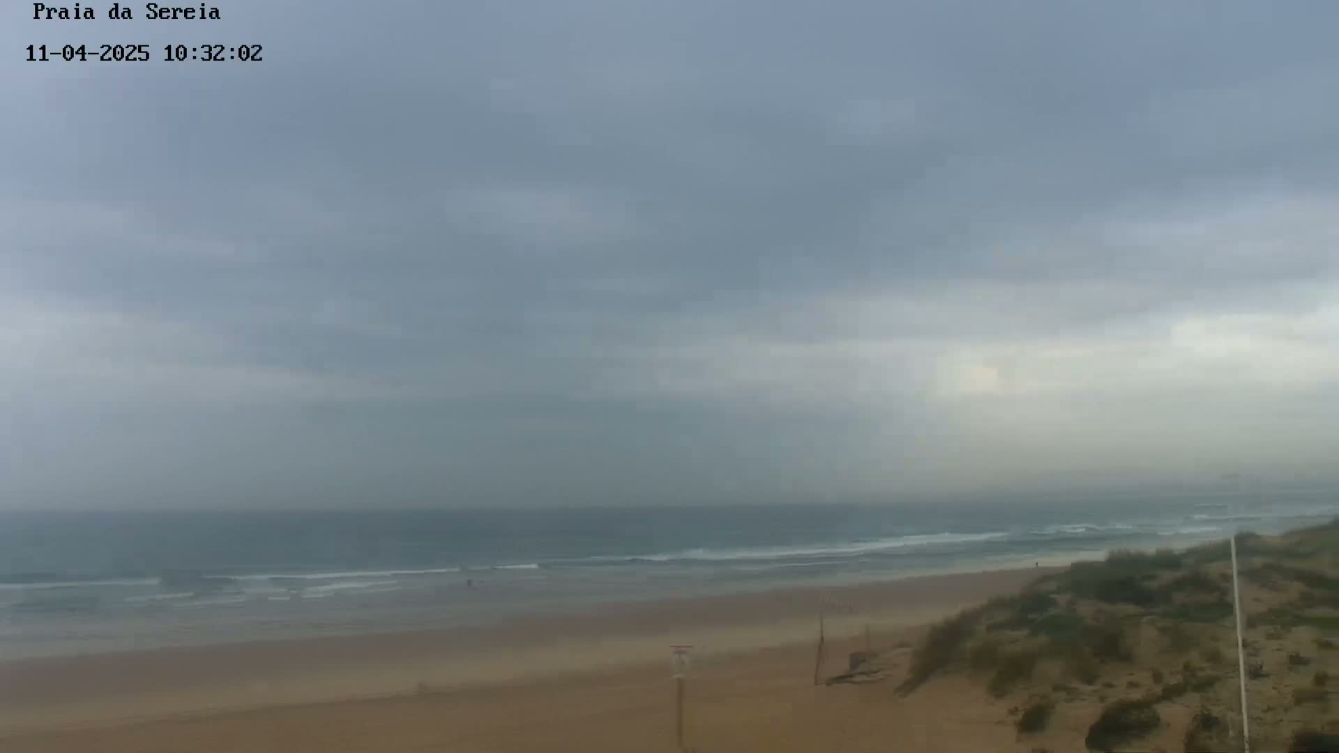 A mostly cloudy day shows a wide sandy beach with small waves lapping the shore, viewed from a slightly elevated dune.
