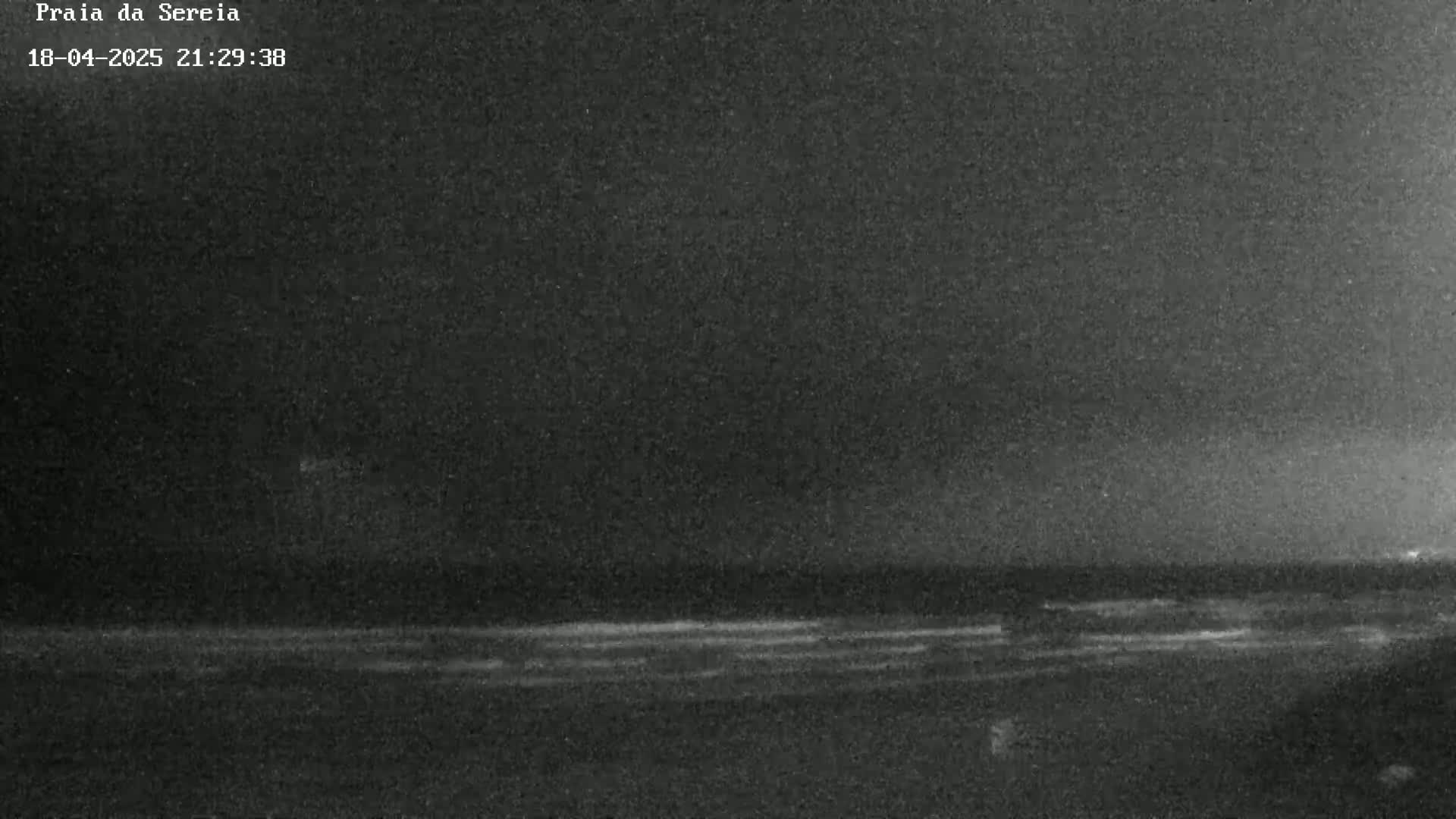 A dark, nighttime image shows a mostly calm ocean with a faint shoreline under a clear sky.