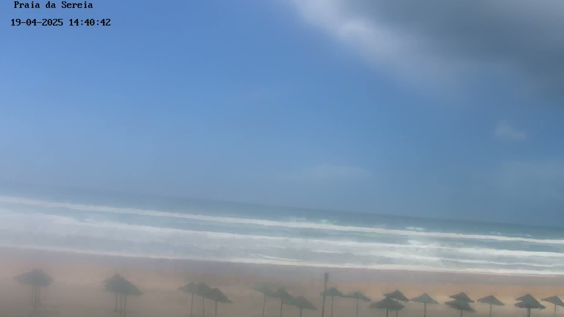 A hazy beach scene shows a sandy shore with several closed umbrellas under a partly cloudy sky and a wavy ocean.