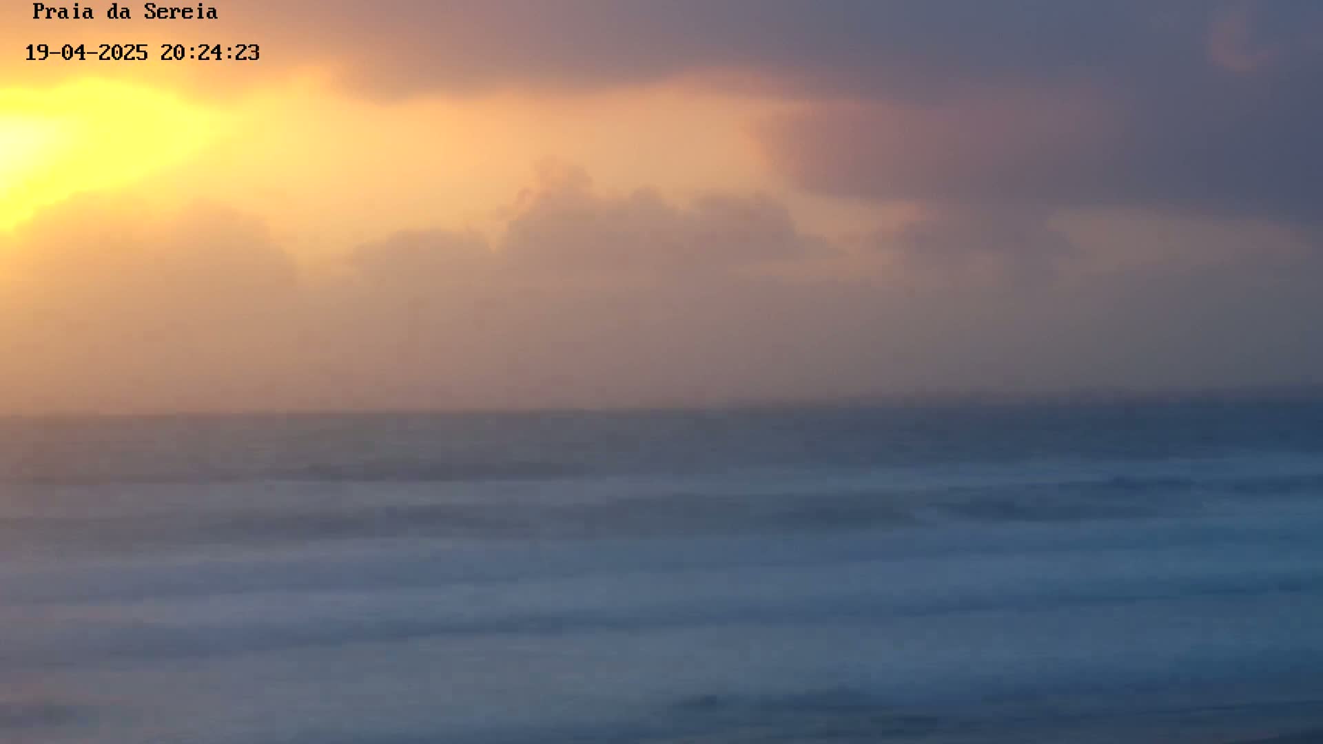 A hazy sunset over a calm ocean with soft, pastel-colored clouds.
