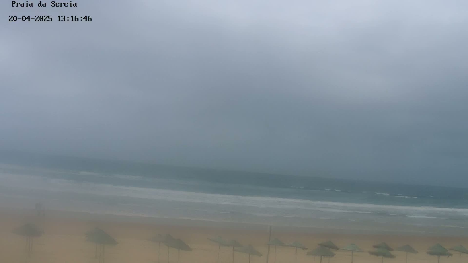 A hazy, overcast sky hangs over a beach with several closed umbrellas and a calm ocean.