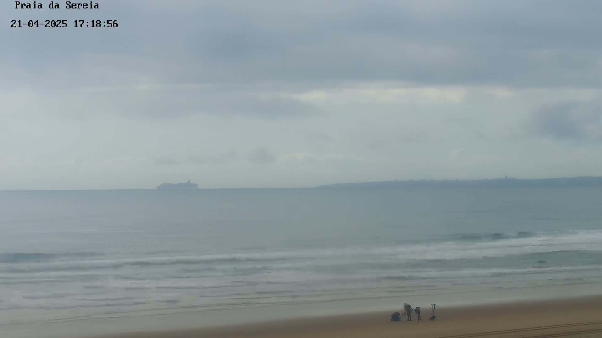 A hazy, overcast day shows a beach with a few people near the water's edge, a distant ship, and a shoreline in the background.