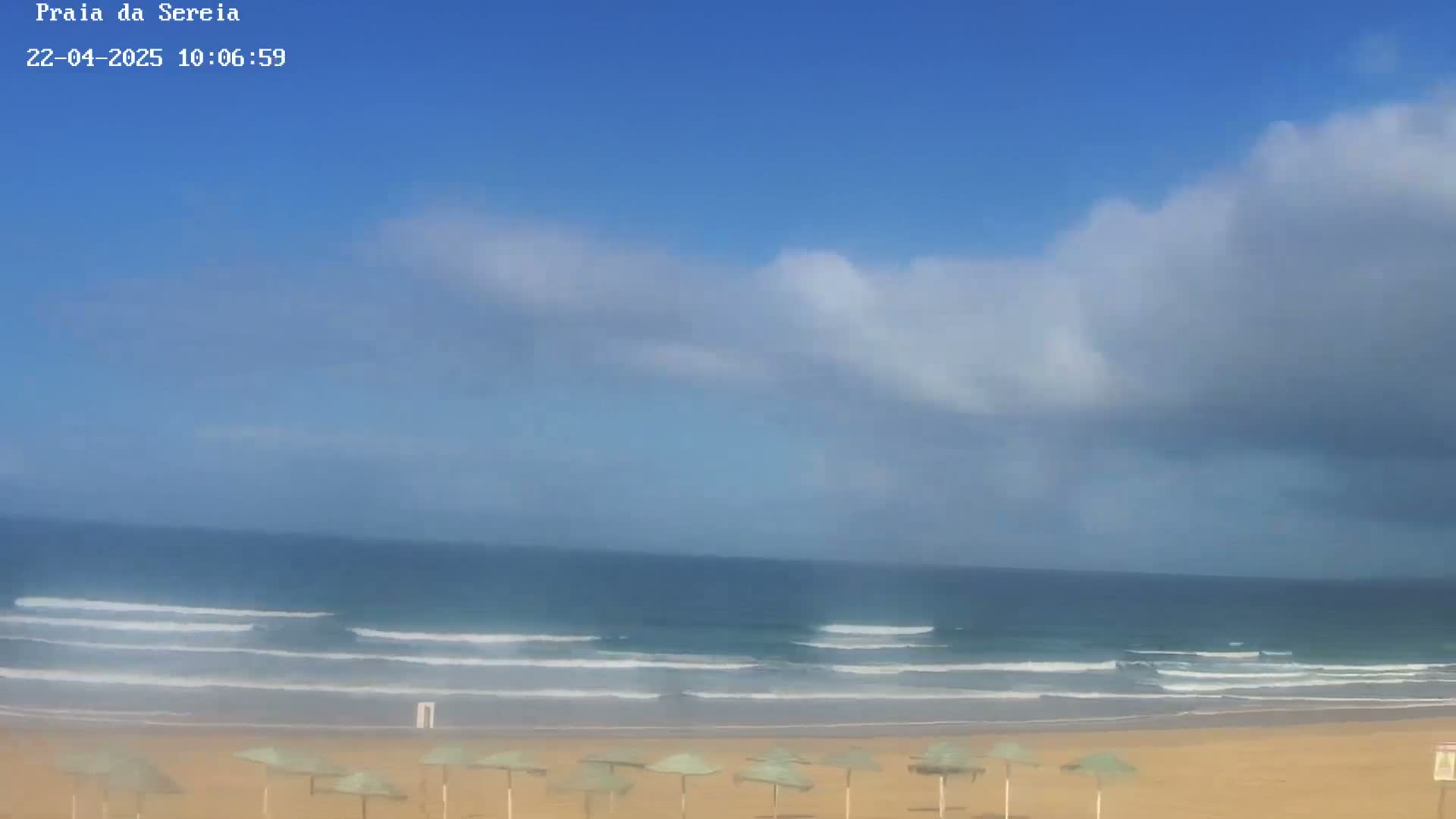 A sandy beach with several closed umbrellas is visible beneath a partly cloudy sky and a calm ocean with small waves.