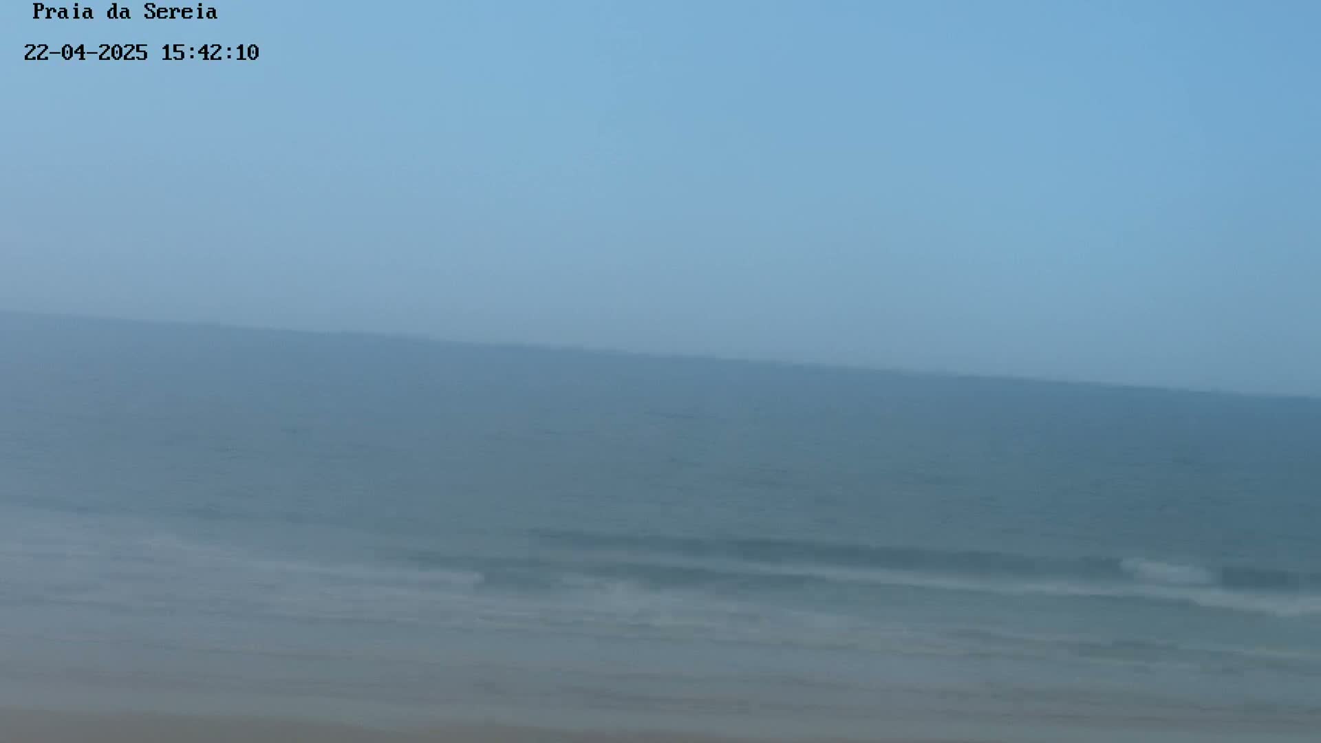 A hazy, overcast sky overlooks a calm ocean with small waves lapping a sandy shore.