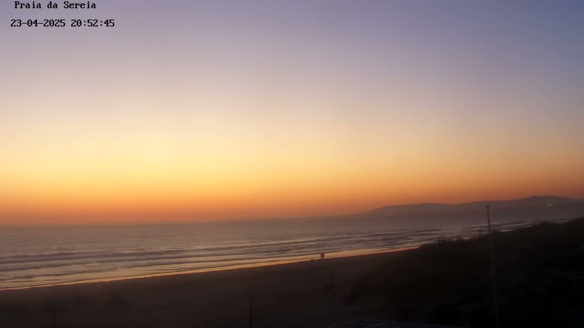An orange and purple sunset over a calm ocean beach with dark sand and distant hills.