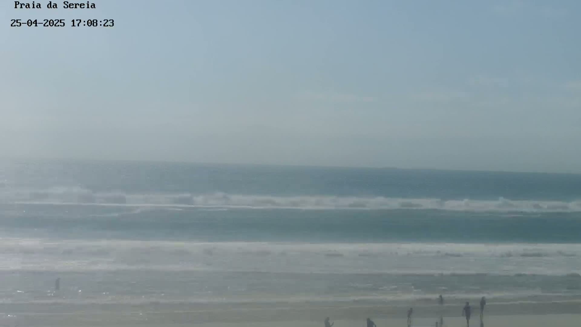A hazy, light-blue ocean with small waves rolls onto a sandy beach where a few people are standing.