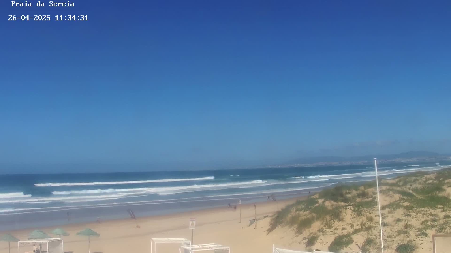 A sandy beach with small waves under a clear blue sky is visible, with sparse beachgoers and sand dunes in the foreground.