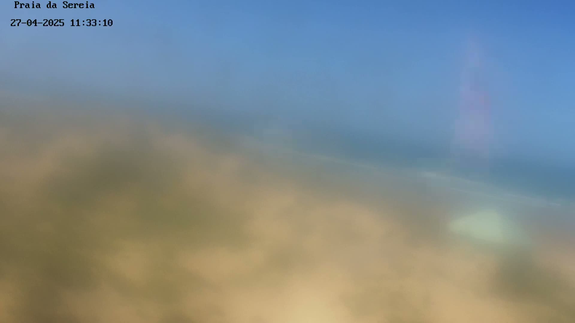 A hazy, blurry view shows a pale sandy beach meeting a muted blue ocean under a clear sky.