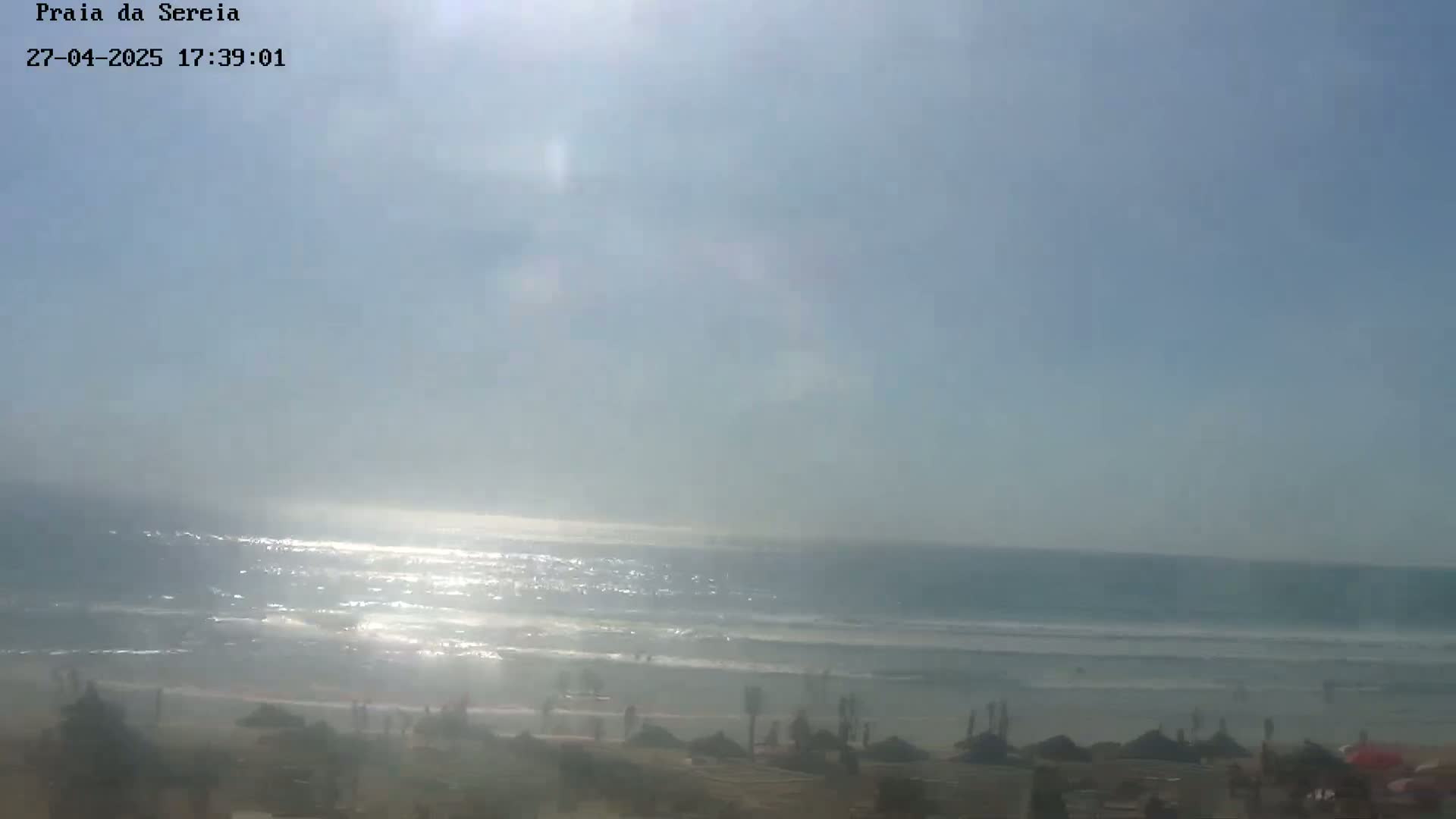 A sunny day at a beach shows a gently waving ocean with some people on the shore and beach umbrellas.