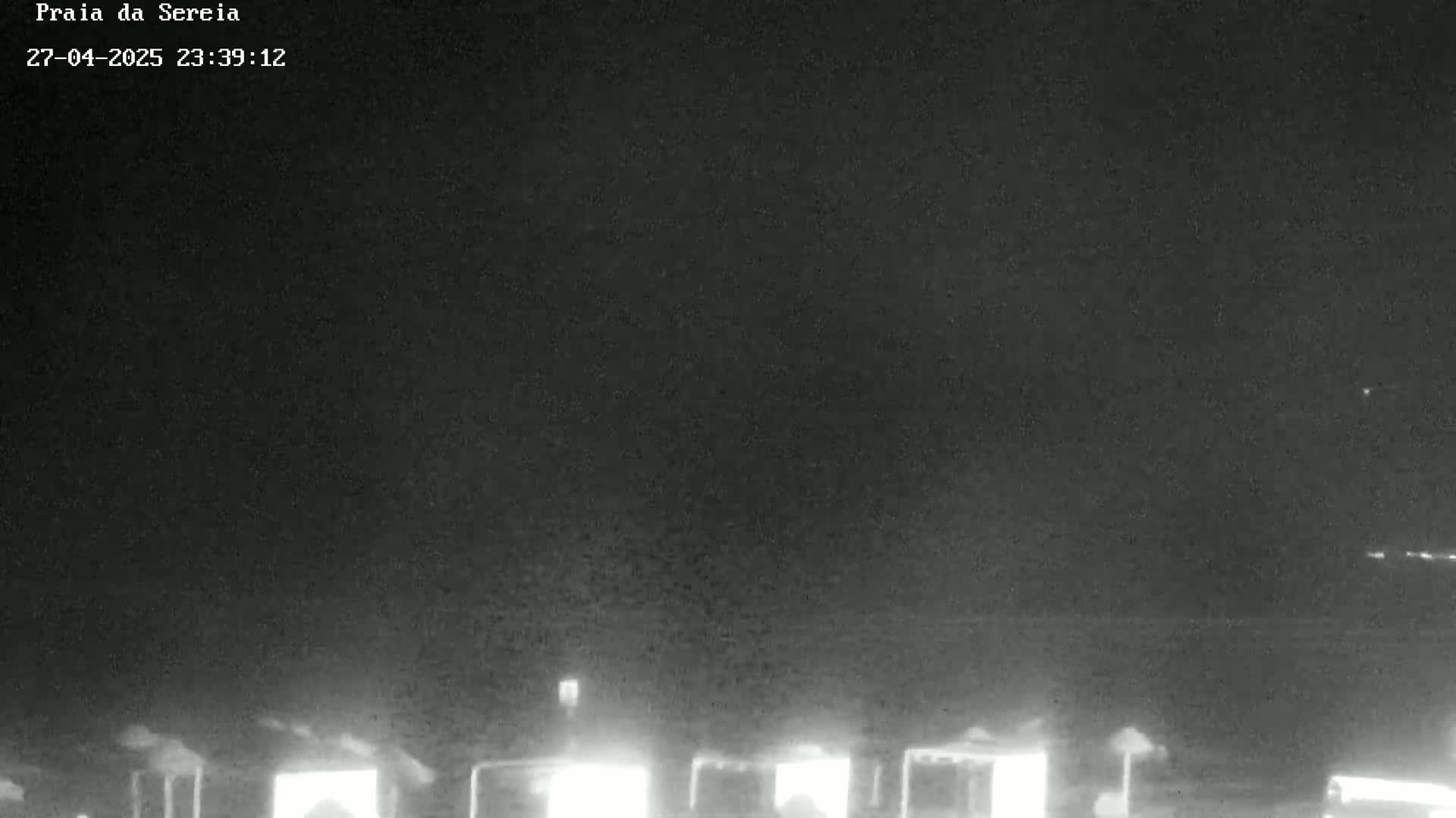 A nighttime, low-light view shows several illuminated structures, possibly beach umbrellas, in the distance under a dark sky.