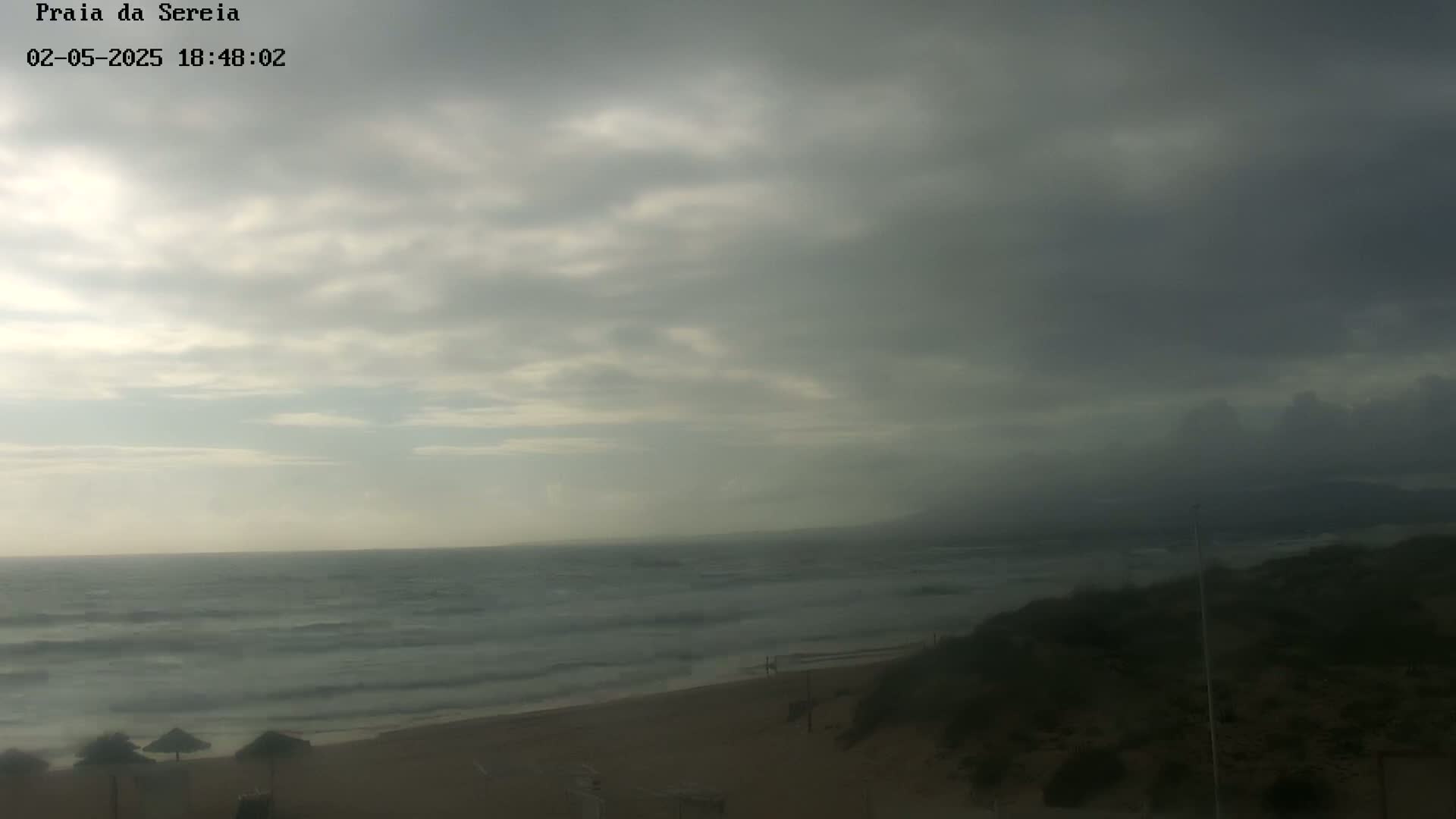 A mostly cloudy, overcast day at a beach shows a somewhat choppy ocean with small waves lapping at a sandy shore, partially obscured by dunes and vegetation.