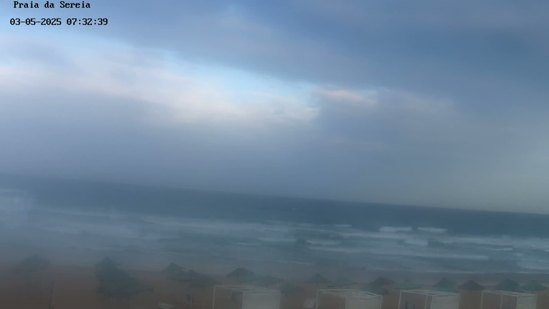 A hazy, overcast sky hangs over a beach with small structures along the shoreline and a wavy ocean in the distance.