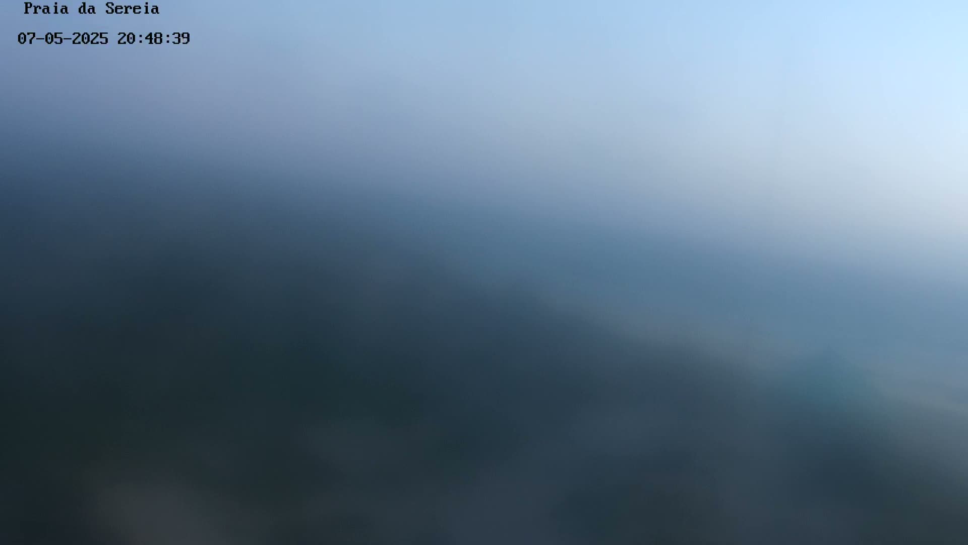 A blurry, hazy image shows a dark, possibly forested landscape under a pale, light-blue sky.