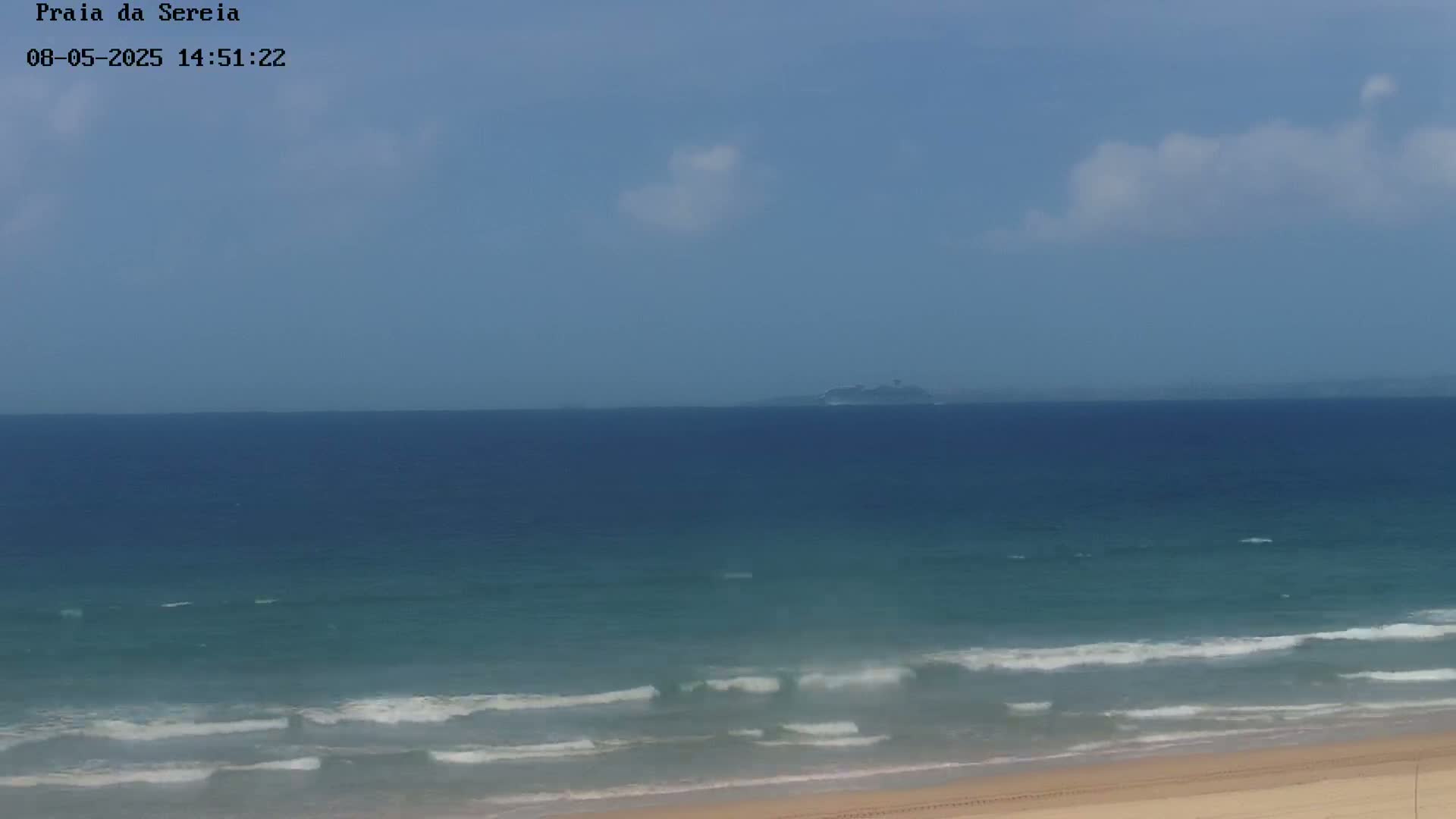 A sandy beach with gentle waves rolls into a calm ocean under a mostly clear, light blue sky with a few scattered clouds.