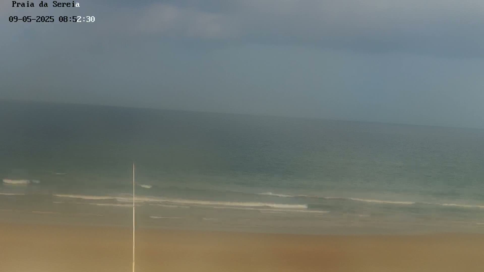 A hazy, overcast day shows a wide sandy beach and calm ocean waves.