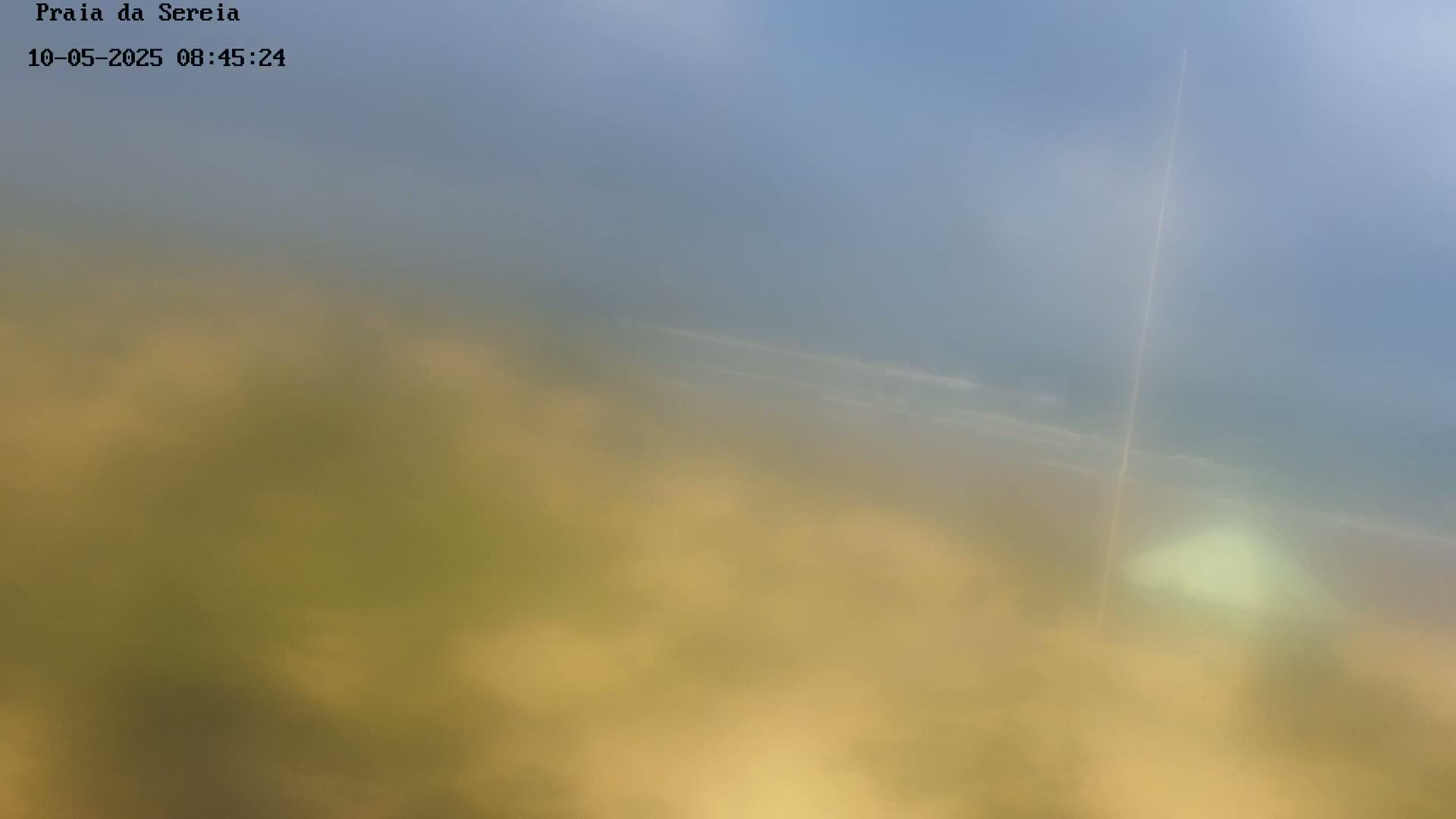 A hazy, blurry view of a beach and ocean under a mostly clear sky shows a faint contrail in the distance.