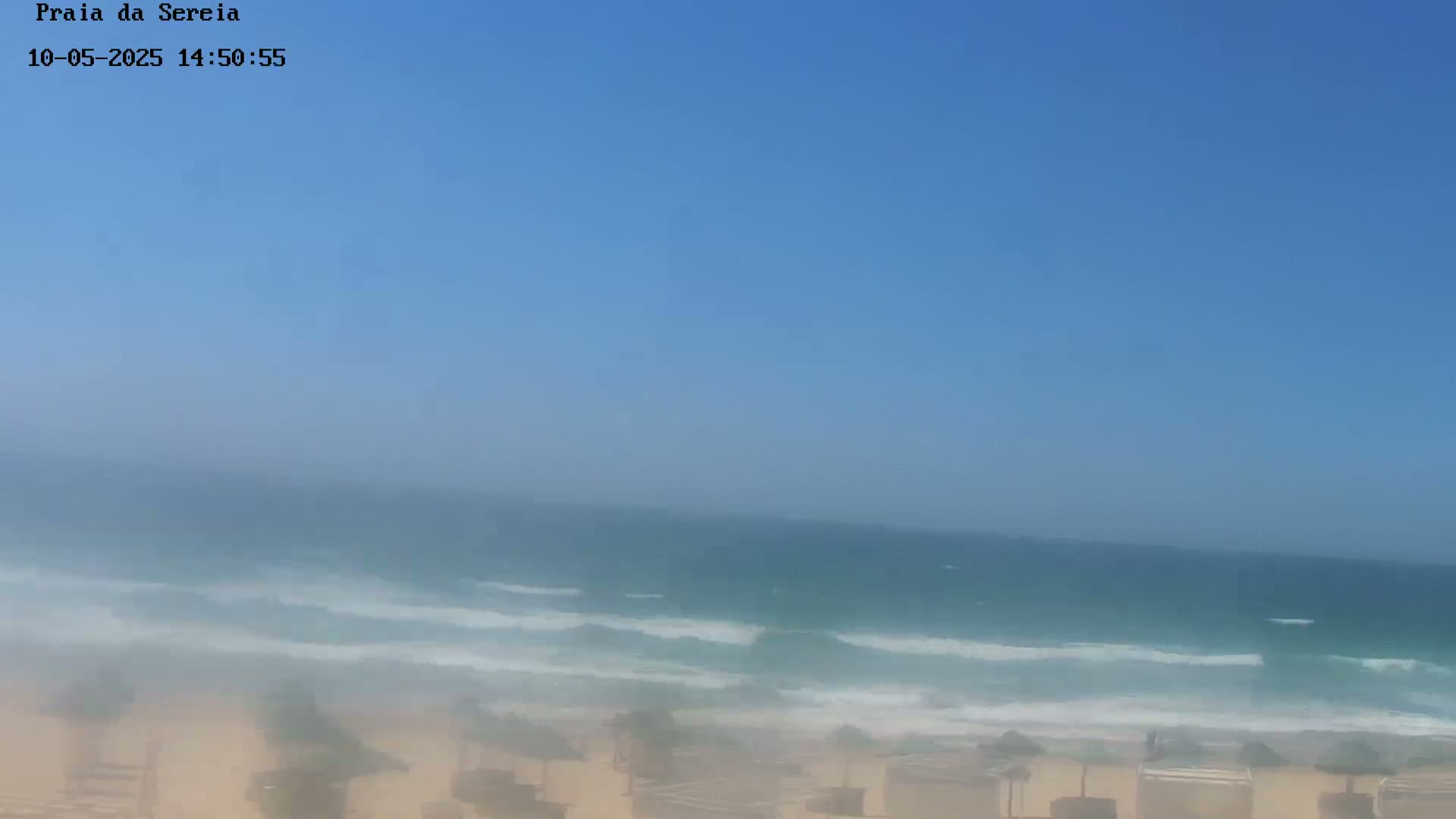 A hazy, sunny day at a beach shows a calm ocean with small waves lapping the shore, and beach umbrellas in the foreground.