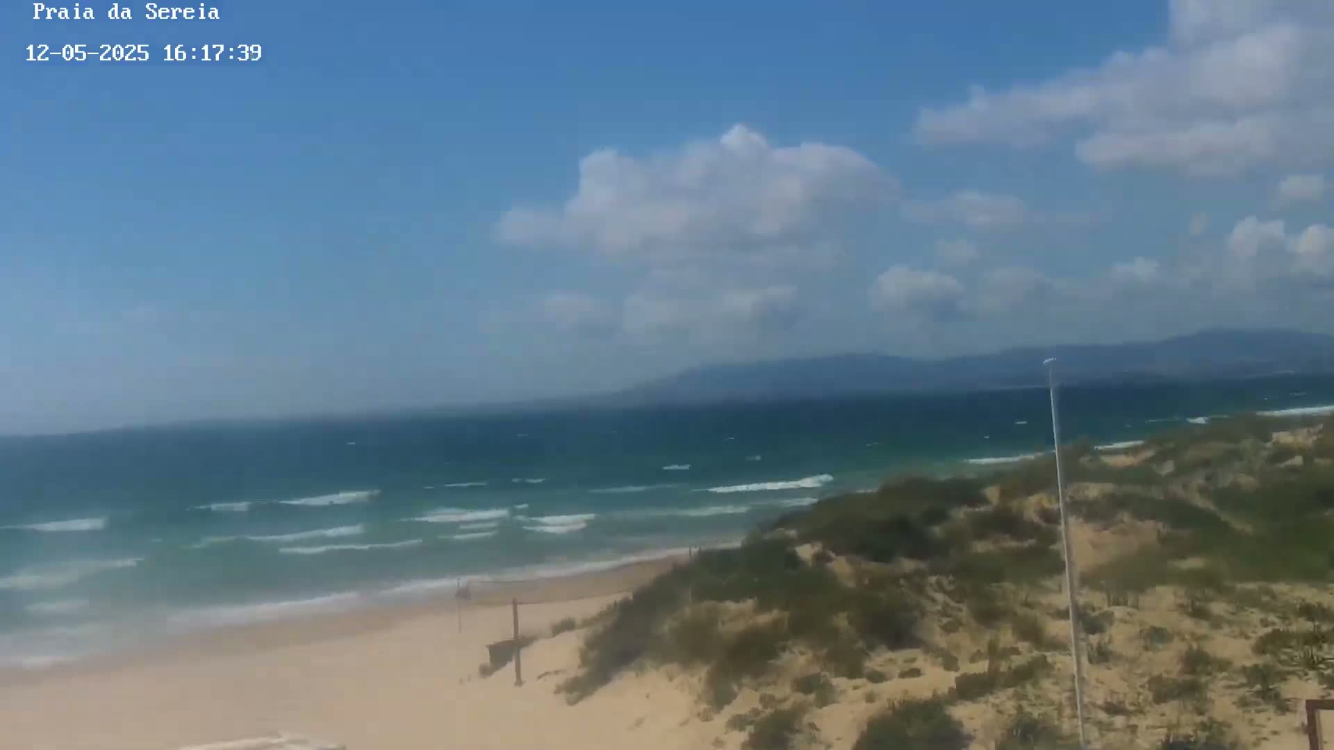 A sandy beach with small waves and partly cloudy skies is viewed from a dune overlooking the ocean and distant mountains.