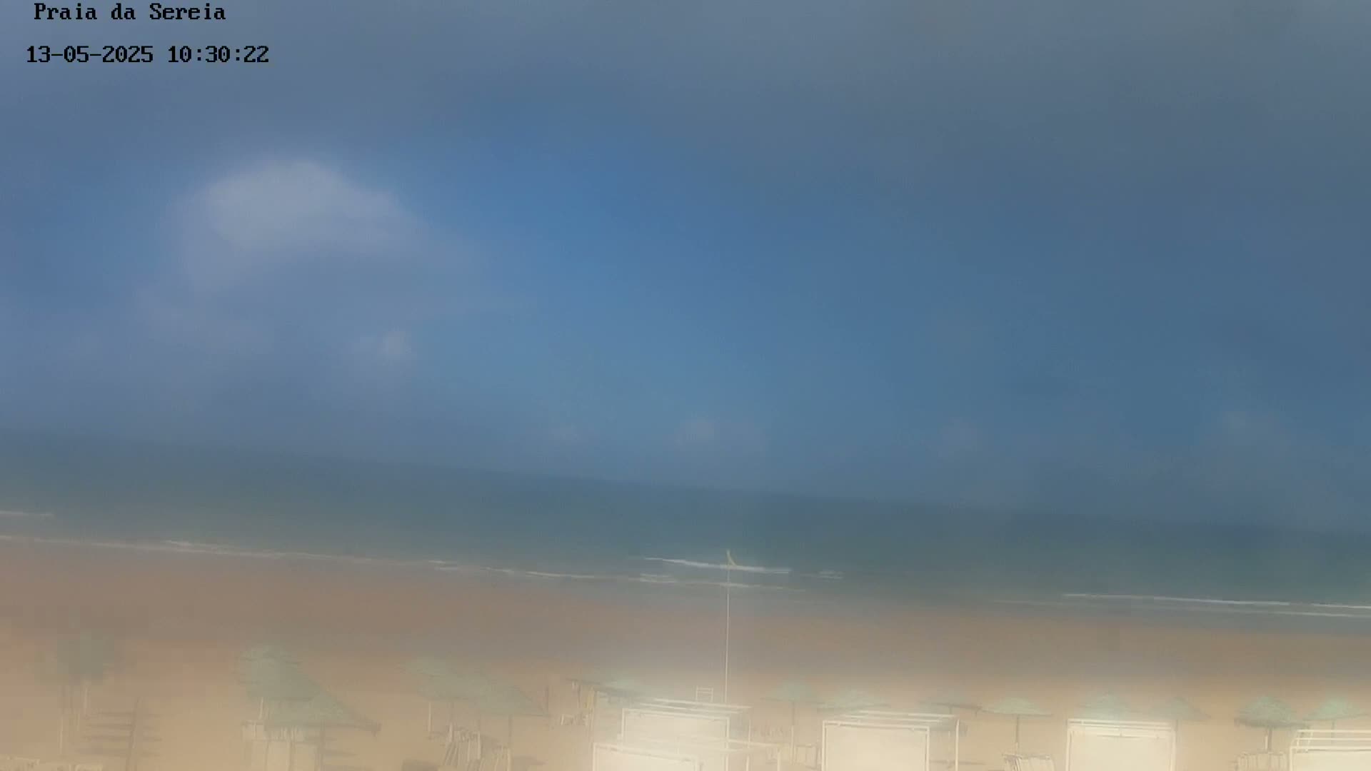 A hazy, mostly clear sky overlooks a calm ocean and sandy beach with rows of beach umbrellas.