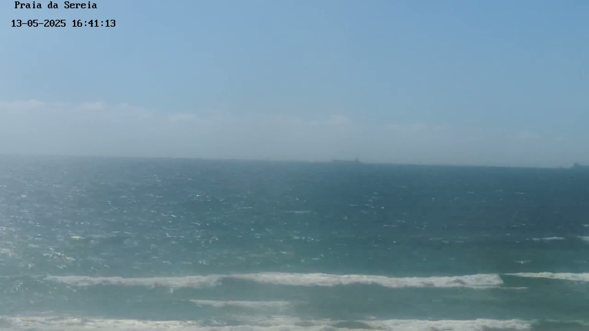 A hazy, sunny day shows a wide expanse of ocean with small waves breaking near the shore.
