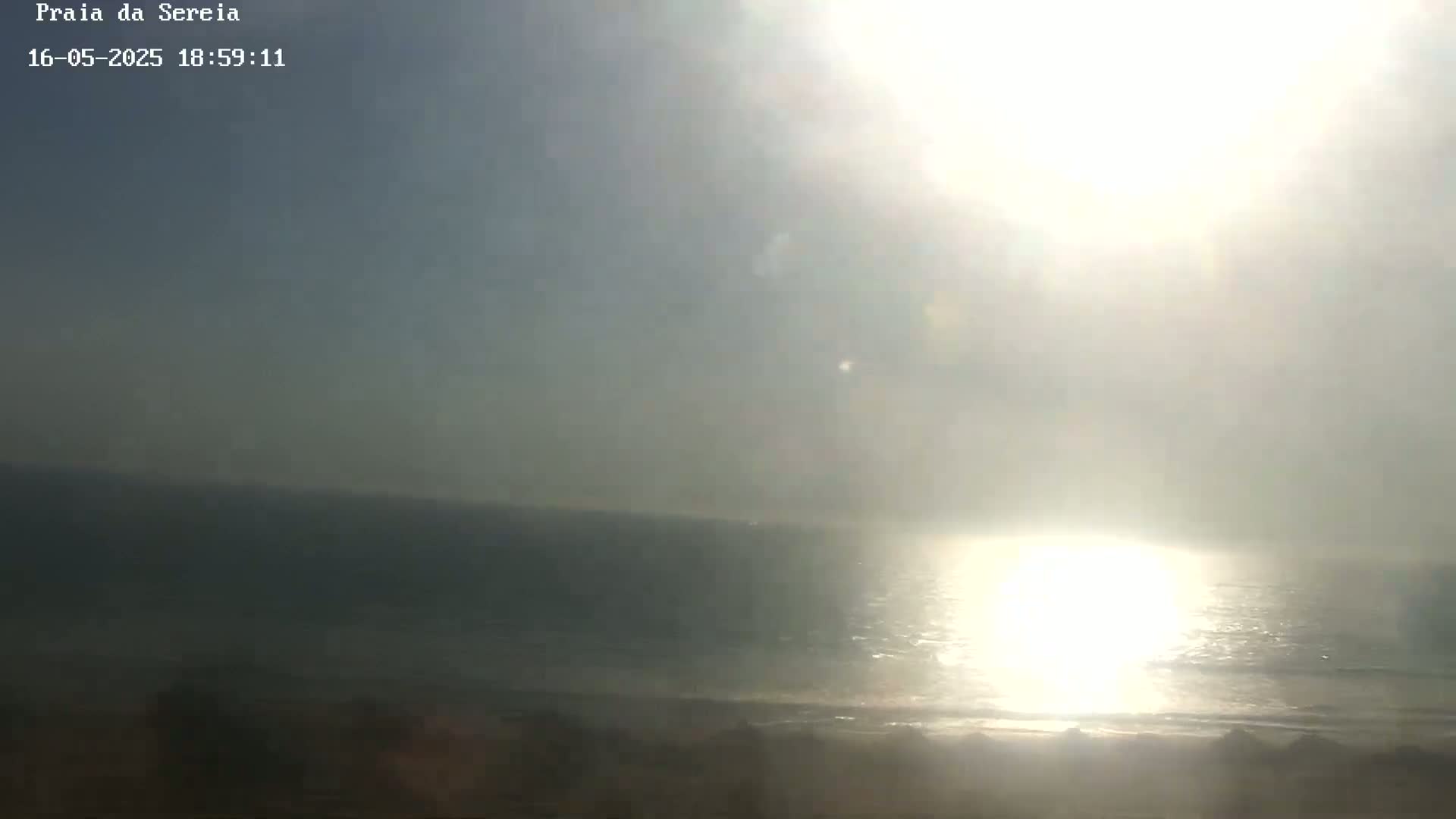 A hazy, bright sun shines over a calm ocean meeting a sandy shore.