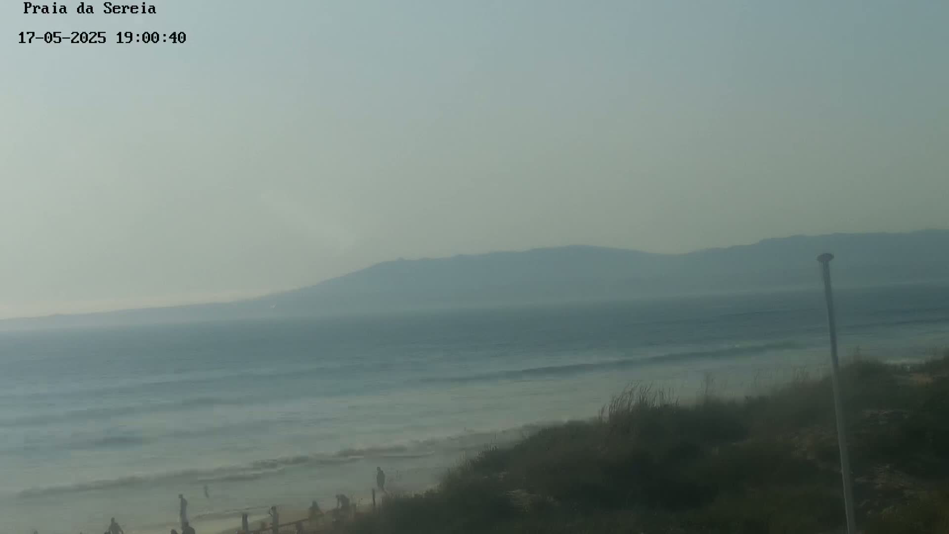 A hazy view of a beach with a calm ocean and distant mountains under a light-grey sky.