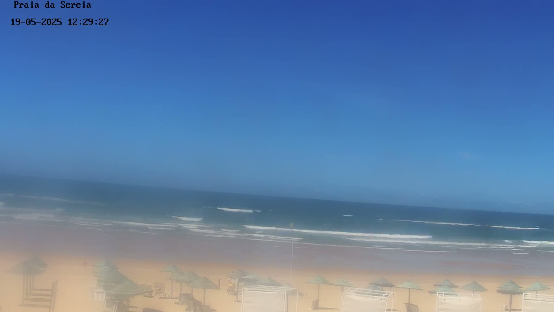A sandy beach with small thatched umbrellas is visible, overlooking a calm ocean under a clear blue sky.