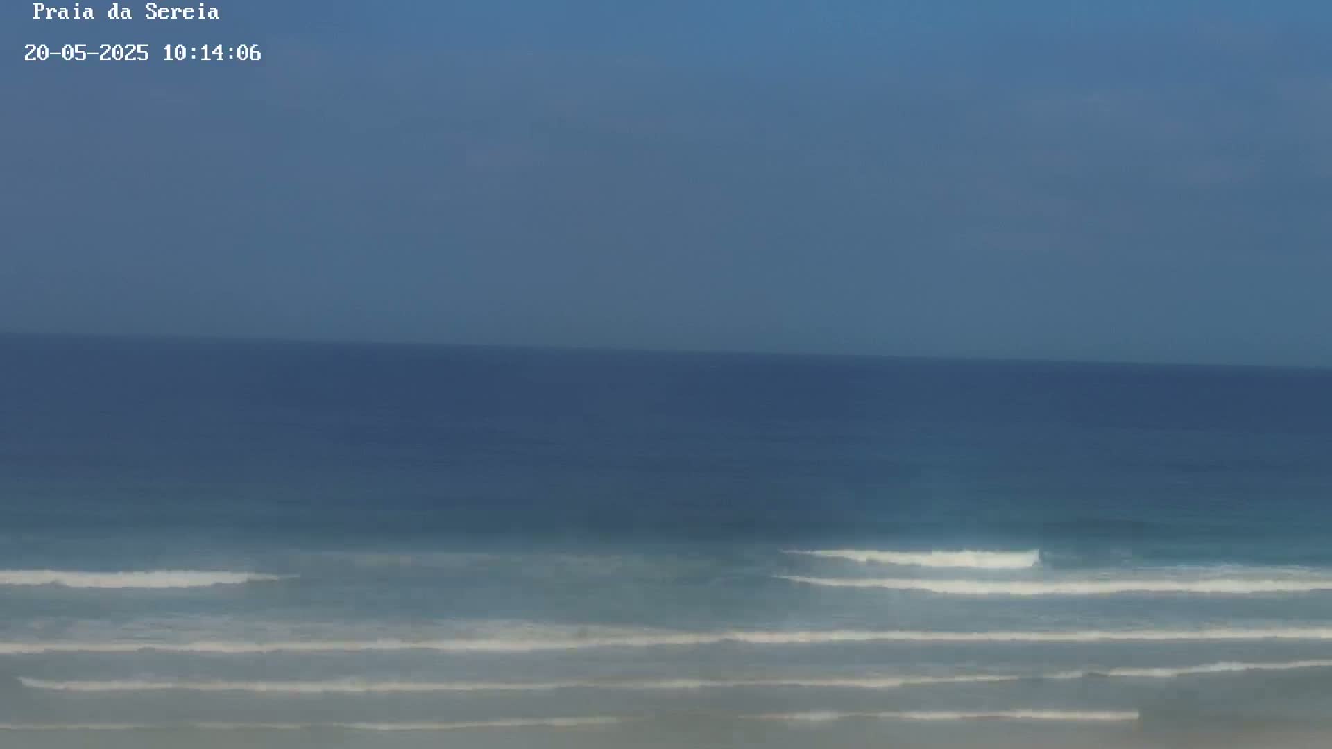A mostly clear, blue sky overlooks a calm ocean with small waves breaking gently on the shore.