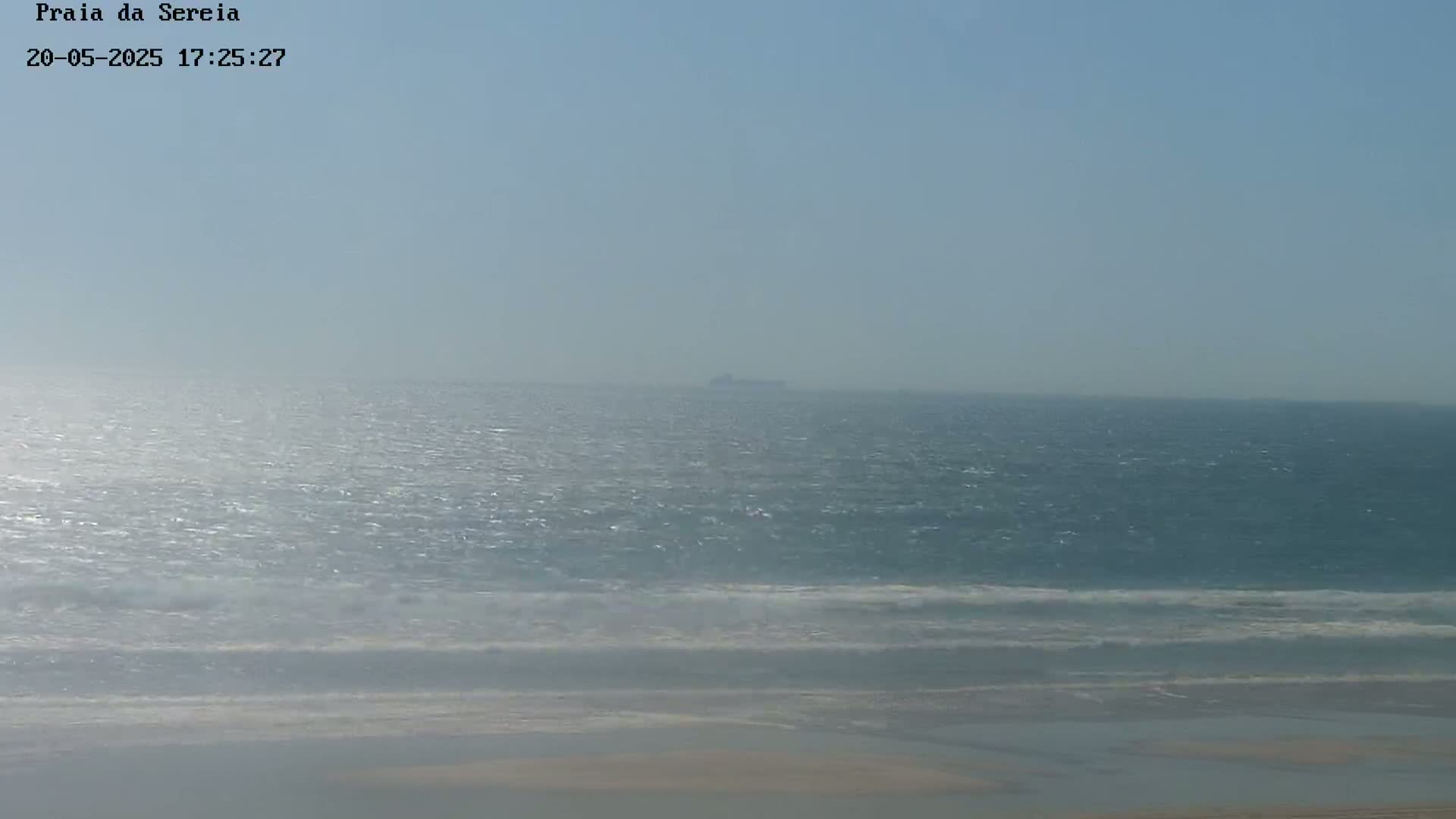 A hazy, sunny day shows a distant ship on a wavy ocean, with a sandy beach in the foreground.