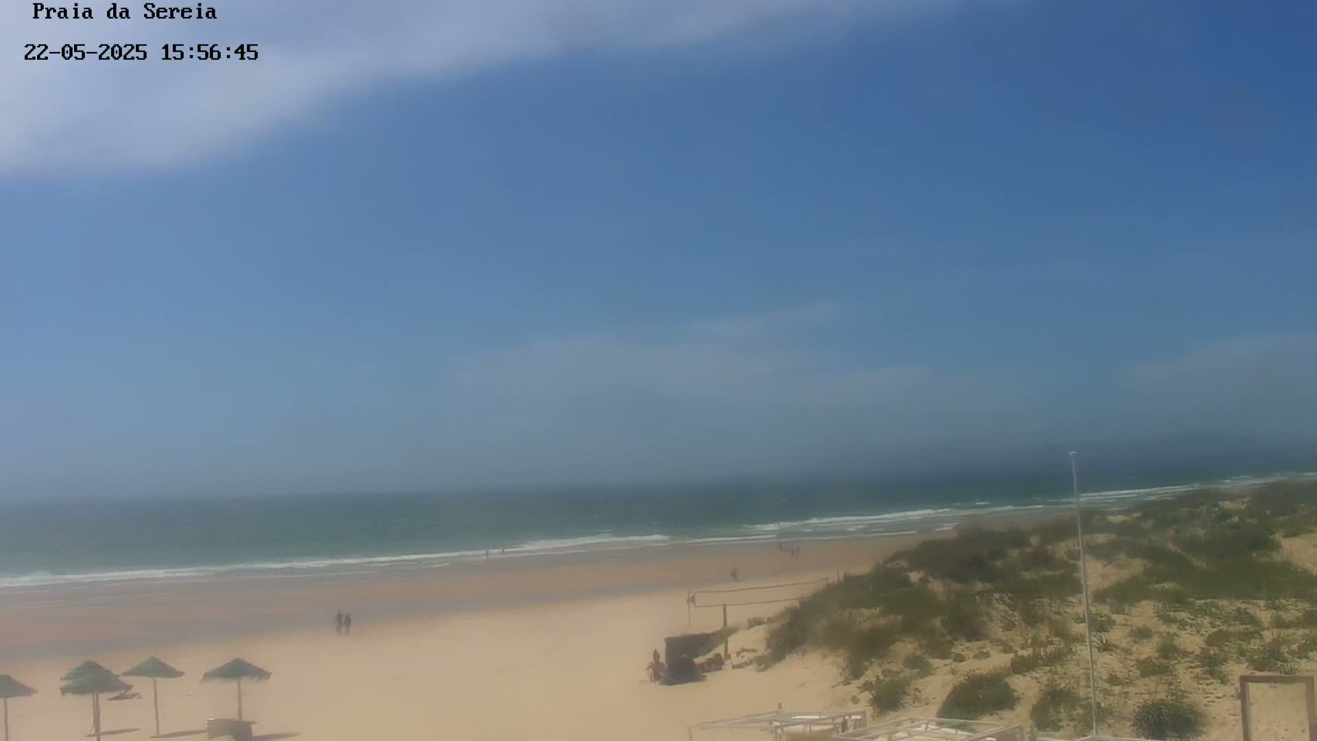 A sandy beach with calm waves under a mostly clear blue sky is visible from a dune-covered vantage point.