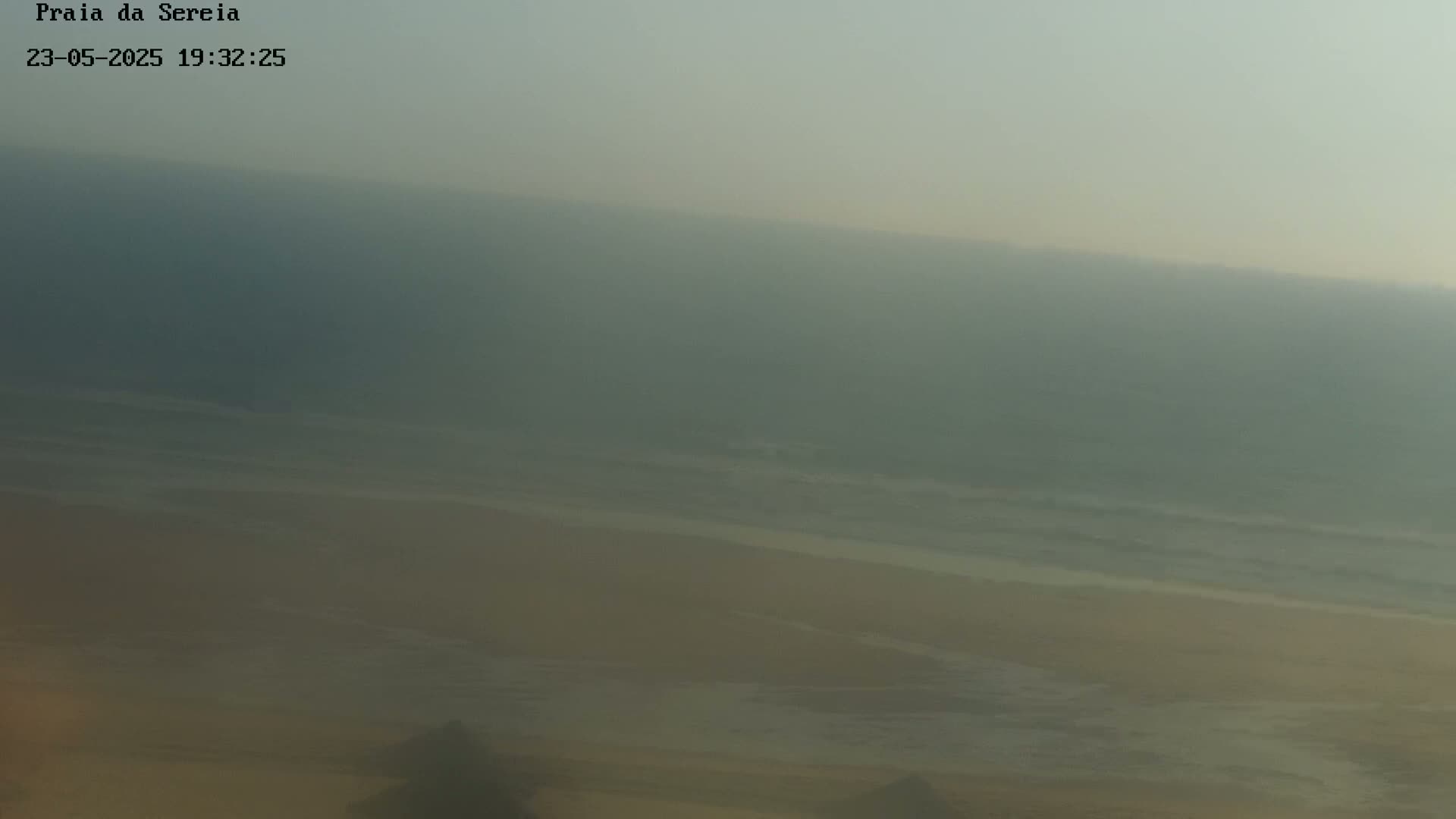 A hazy, overcast view shows a sandy beach meeting a muted ocean with small waves.