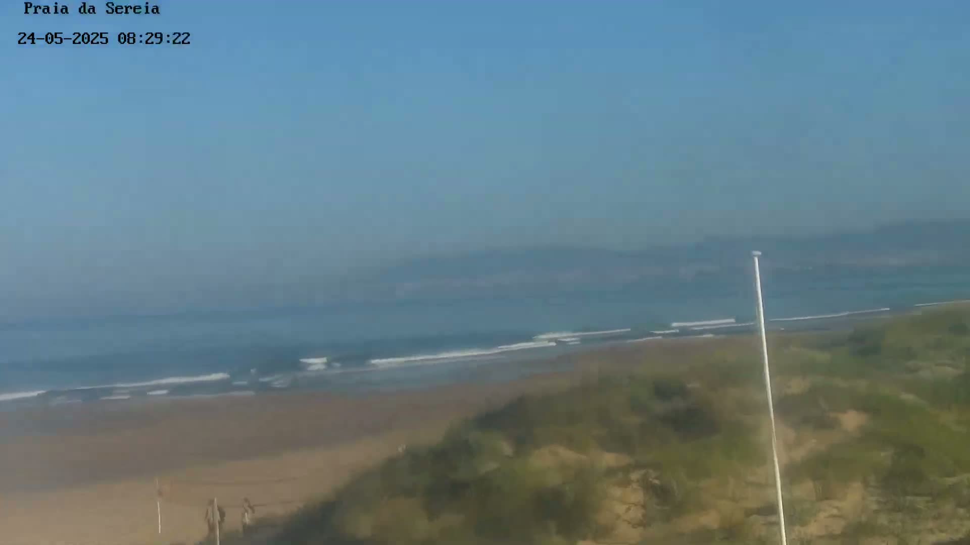 A hazy, sunny day overlooks a sandy beach with gentle waves lapping the shore, backed by low-lying vegetation and distant hills.