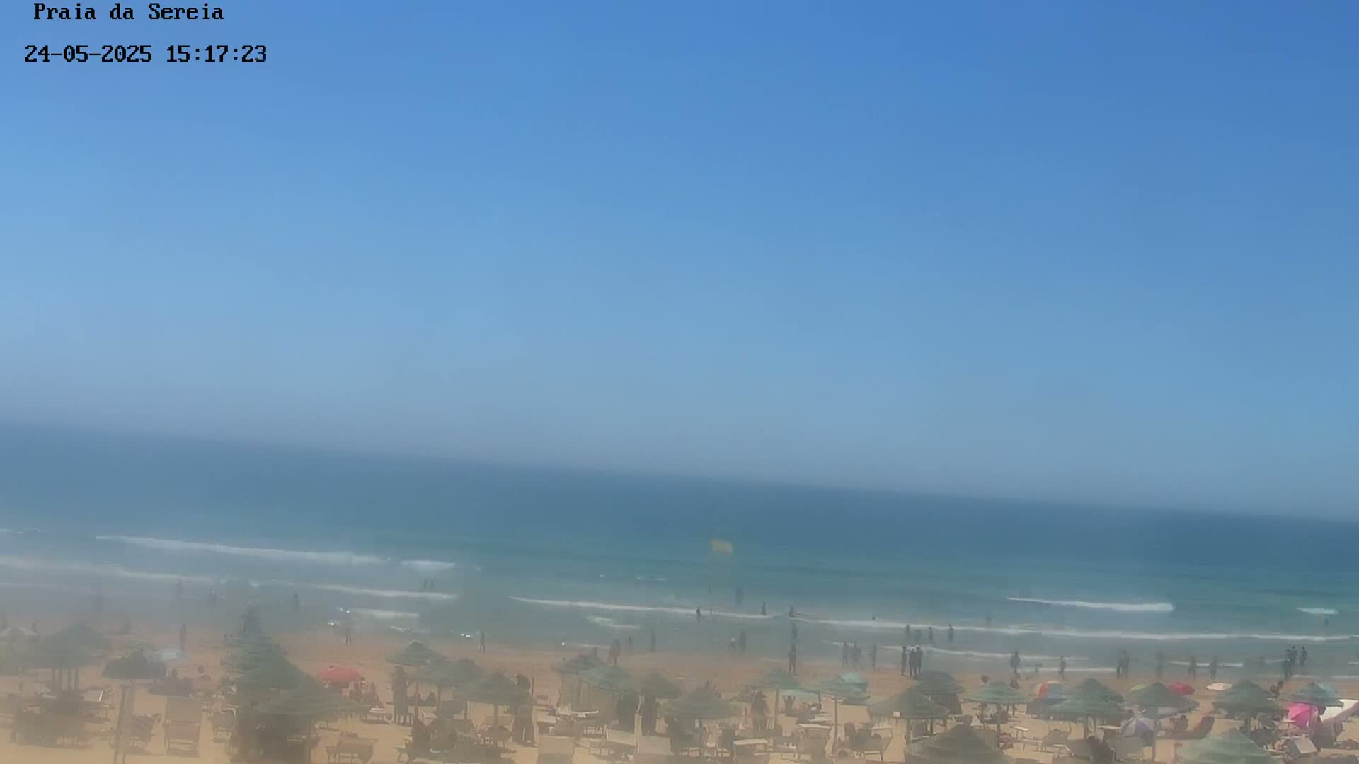 A hazy beach scene shows a calm ocean with a sandy shore dotted with beach umbrellas and people under a clear blue sky.