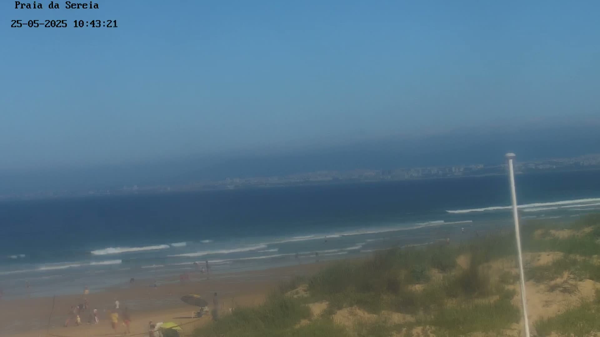 A sandy beach with small waves and a few people on it stretches toward a hazy city skyline under a clear blue sky.