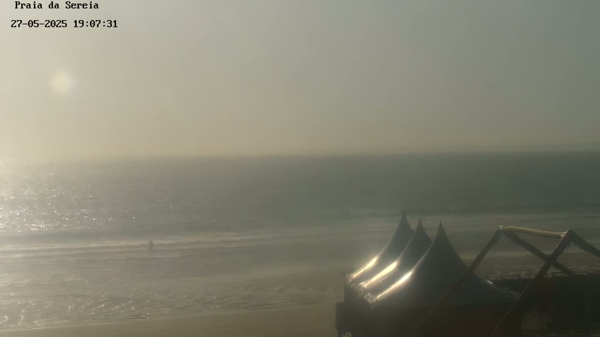 A hazy, sunlit beach scene shows a calm ocean with a lone person in the water and three dark, conical tents on the sand.