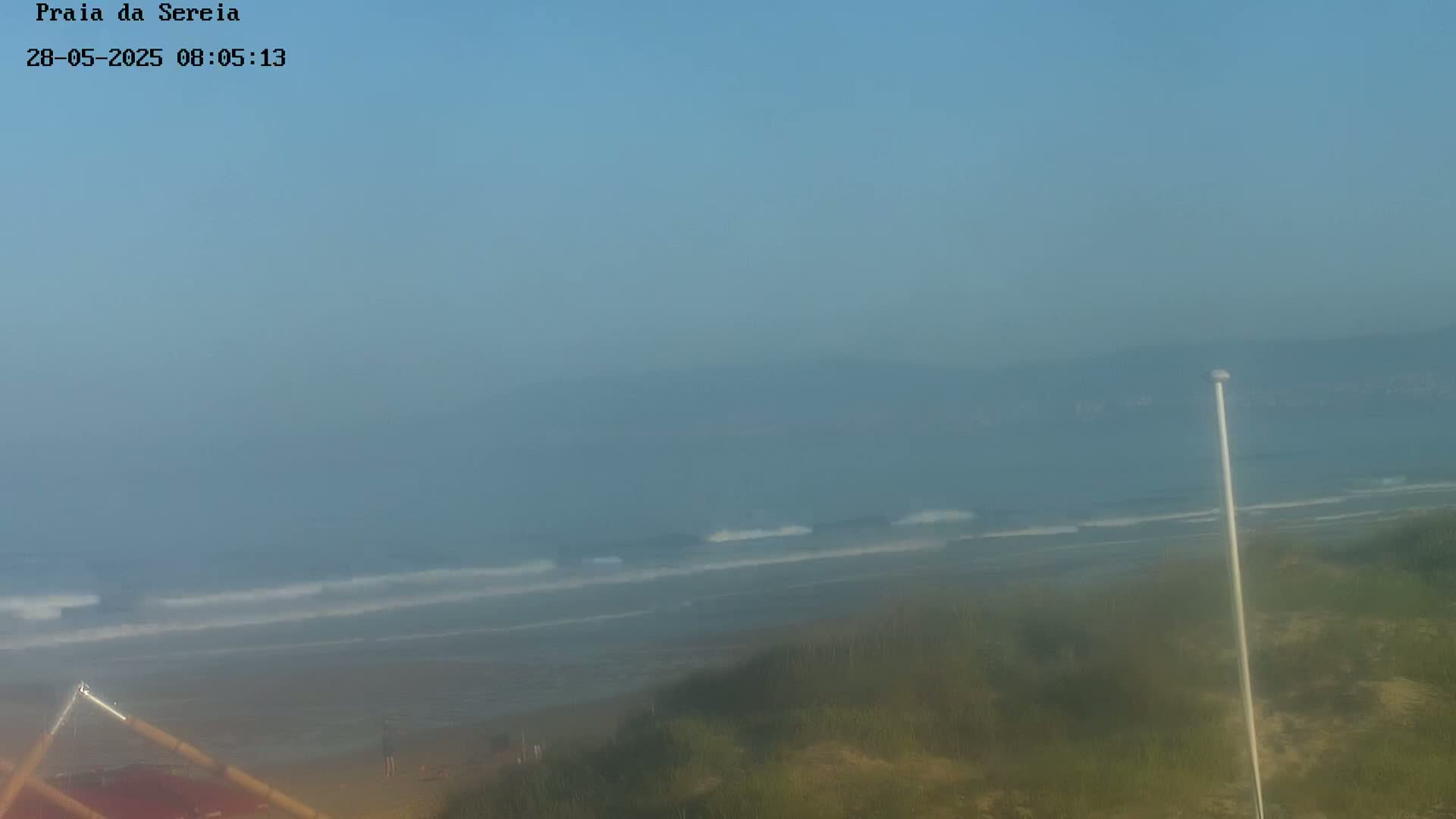 A hazy view shows a beach with small waves gently rolling onto the shore, under a clear, light-blue sky.